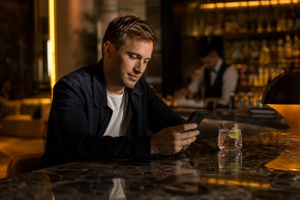 Man in a navy suit jacket checking his phone at a hotel bar counter in the evening, illustrative photo, not a member