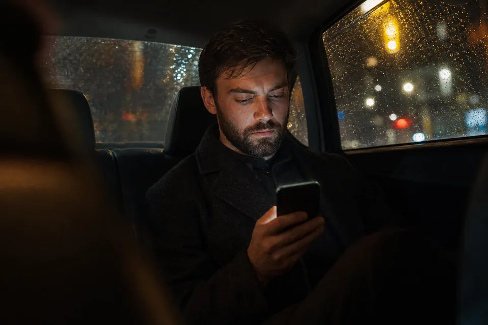 Man scrolling profiles on his phone in the back seat of a taxi at night, city lights outside, illustrative photo, not a member