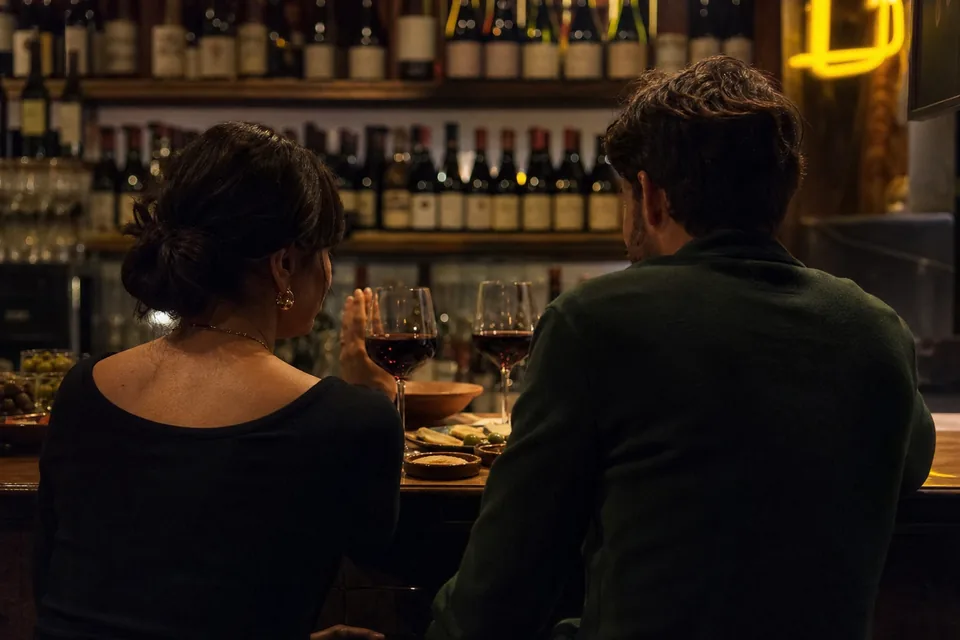 Couple sharing small plates at a wine bar counter at night, seen from behind, illustrative photo, not a member